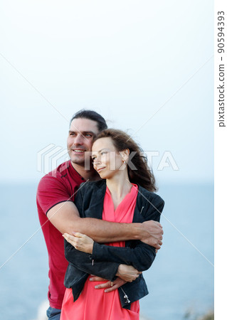 Portrait of lovely middle-aged couple looking aside at copyspace and hugging while standing at the seaside rock cliff edge. 90594393