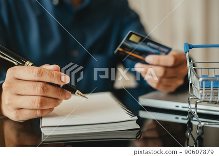 Business men sits at his desk at home holding a credit card and writing a summary of expenses in a notebook. 90607879
