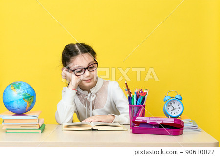 The schoolgirl sits at the desk and thinks about the decision of the task on yellow background. 90610022