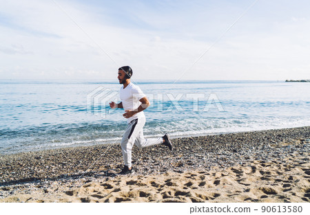 Confident young ethnic sportsman jogging on seashore Confident young ethnic sportsman jogging on seashore 90613580