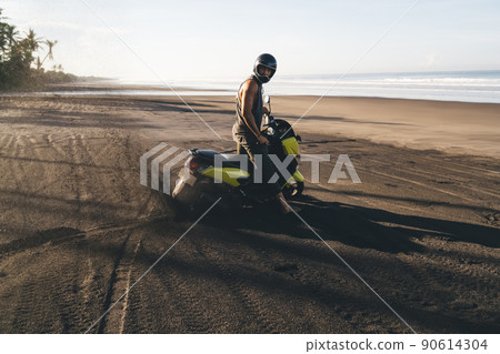 Sporty biker standing with motorbike on sandy coastline Sporty biker standing with motorbike on sandy coastline 90614304