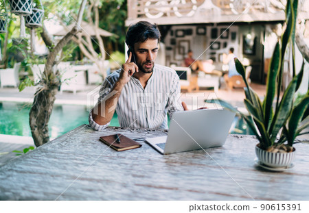 Ethnic man talking on smartphone and working on laptop remotely 90615391