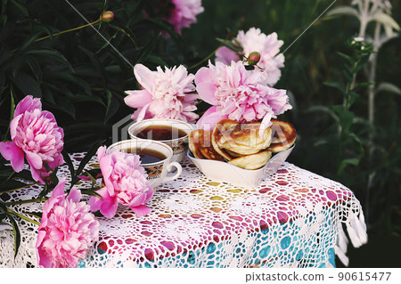 Tea in country style in summer garden. Two cups of black tea and pancakes on handmade crocheted vintage lacy tablecloth and blooming peony flowers in sunlight. Tea in country style in summer garden. Two cups of black tea and pancakes on handmade crocheted vintage lacy tablecloth and blooming peony flowers in sunlight. 90615477