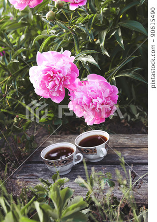 Tea in country style in summer garden in the village. Two cups of black tea on wooden boards and blooming peony flowers in sunlight. Tea in country style in summer garden in the village. Two cups of black tea on wooden boards and blooming peony flowers in sunlight. 90615490