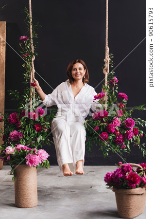 attractive and happy woman in white cozy clothes on a swing with peony flowers 90615793