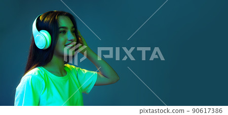 Portrait of attractive young girl with long loose hair listening to music isolated on blue background in neon light. Concept of beauty, art, fashion, emotions. Flyer Portrait of attractive young girl with long loose hair listening to music isolated on blue background in neon light. Concept of beauty, art, fashion, emotions. Flyer 90617386