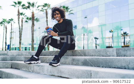 Cheerful young sportsman sitting on stairs with bottle of water Cheerful young sportsman sitting on stairs with bottle of water 90617522