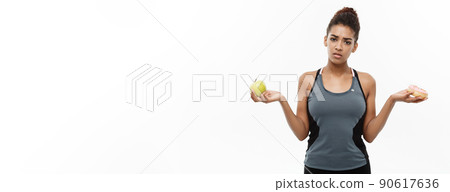 Healthy and diet concept - Beautiful sporty African American make a decision between donut and green apple. Isolated on white background. Healthy and diet concept - Beautiful sporty African American make a decision between donut and green apple. Isolated on white background. 90617636