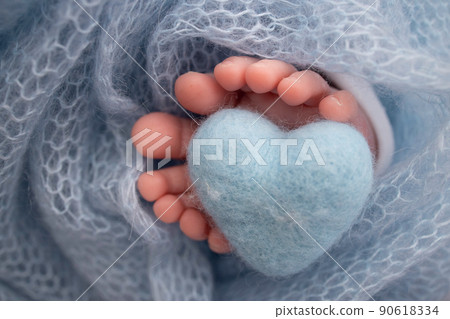 Knitted blue heart in the legs of a baby. Soft feet of a new born in a blue wool blanket. Close-up of toes, heels and feet of a newborn. Macro photography the tiny foot of a newborn baby. Knitted blue heart in the legs of a baby. Soft feet of a new born in a blue wool blanket. Close-up of toes, heels and feet of a newborn. Macro photography the tiny foot of a newborn baby. 90618334