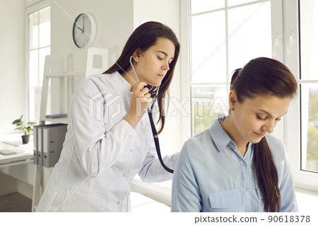 Young female doctor checks woman's breathing and heartbeat at appointment at medical clinic. 90618378