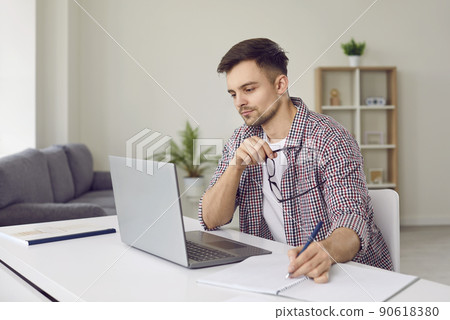 Concentrated young man listens to online lecture and writes information in notebook. Concentrated young man listens to online lecture and writes information in notebook. 90618380