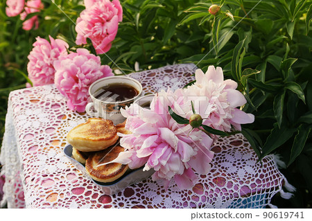 Tea in country style in summer garden. Two cups of black tea and pancakes on handmade crocheted vintage lacy tablecloth and blooming peony flowers in sunlight. 90619741