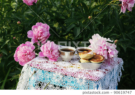 Tea in country style in summer garden. Two cups of black tea and pancakes on handmade crocheted vintage lacy tablecloth and blooming peony flowers in sunlight. Tea in country style in summer garden. Two cups of black tea and pancakes on handmade crocheted vintage lacy tablecloth and blooming peony flowers in sunlight. 90619747