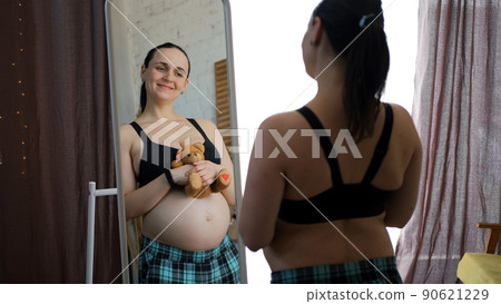 Happy Pregnant Woman Touching her Belly while Holding Teddy Bear in Hands in Front of the Mirror Indoors. Expectation of a Baby Concept Happy Pregnant Woman Touching her Belly while Holding Teddy Bear in Hands in Front of the Mirror Indoors. Expectation of a Baby Concept 90621229