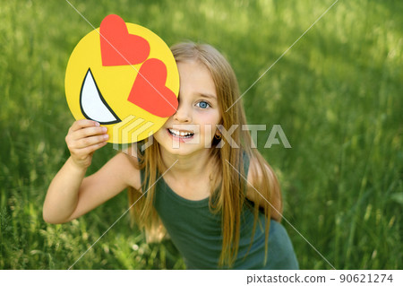 Sweet blue-eyed little girl is holding a cardboard love emoticon with hearts in hands. A happy child on a natural background 90621274