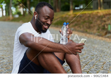 Young adult man resting on a park after a run with a bottle of water and smartphone. Outdoor workout 90621464