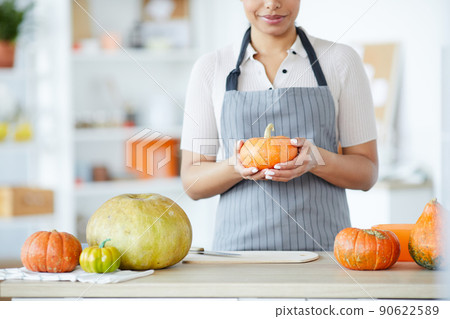 Close-up of skilled woman in apron standing at counter and choosing pumpkin to make jack-o-lantern 90622589