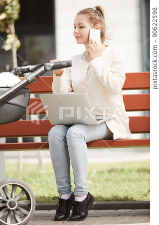 Young mother in white coat working outside on her smartphone and laptop while baby sleeping in stroller 90622598