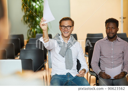 Cheerful excited young hipster man in eyeglasses sitting in auditorium and raising hand with paper while showing it to speaker 90623562