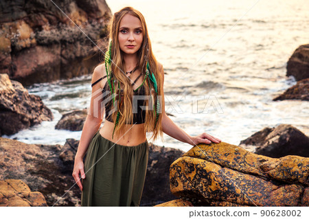 portrait of stylish elegant redheaded chestnut blonde young girl in indie bohemian bo-ho style clothes black stylish top.boho woman long hair,makeup and green feathers in head on the rocks by the sea 90628002