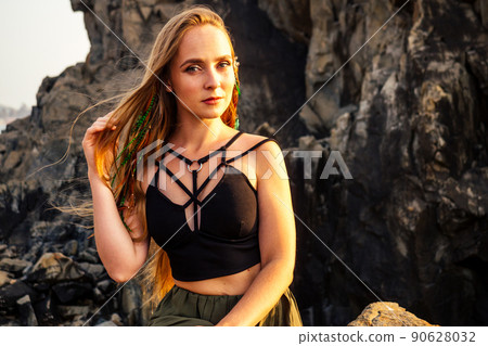 portrait of stylish elegant redheaded chestnut blonde young girl in indie bohemian bo-ho style clothes black stylish top.boho woman long hair,makeup and green feathers in head on the rocks by the sea 90628032