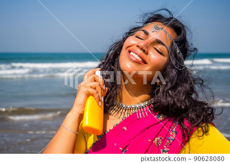 portrait of a beautiful smiling snow-white smile indian woman black curly hair and dark skin in a pink sari holding bottle of sunscreen spray on beach.girl enjoying spf body paradise vacation 90628080