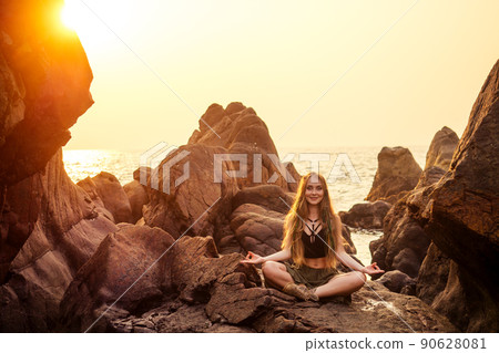 stylish redheaded chestnut blonde young girl in indie bohemian bo-ho style clothes black stylish top.boho woman long hair,makeup and green feathers in head practicing yoga asanas on the rocks by sea 90628081