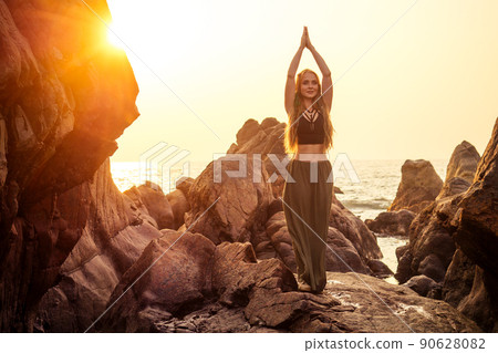stylish redheaded chestnut blonde young girl in indie bohemian bo-ho style clothes black stylish top.boho woman long hair,makeup and green feathers in head practicing yoga asanas on the rocks by sea 90628082