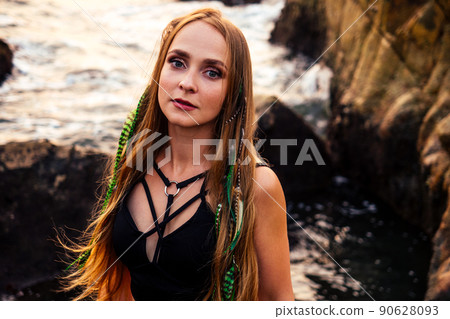 portrait of stylish elegant redheaded chestnut blonde young girl in indie bohemian bo-ho style clothes black stylish top.boho woman long hair,makeup and green feathers in head on the rocks by the sea 90628093