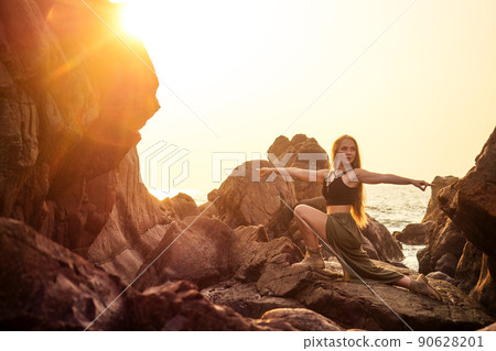 stylish redheaded chestnut blonde young girl in indie bohemian bo-ho style clothes black stylish top.boho woman long hair,makeup and green feathers in head practicing yoga asanas on the rocks by sea 90628201