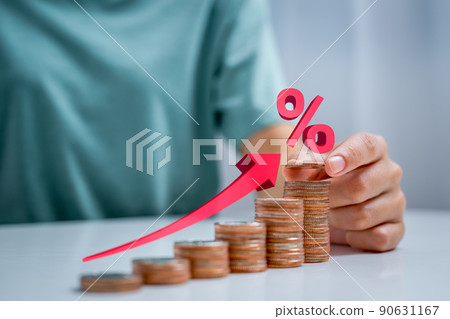 Woman's hand placing coin on stack of coins arranged in upward position and illustration of red arrow pointing upward indicating rising interest rates. 90631167