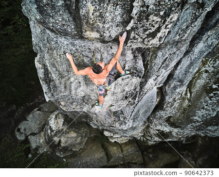 Young man climber climbing difficult route on a high rock with rope. Fearless rock climber training rock climbing on summer day. Concept of extreme sport, adventures and active lifestyle. Aerial view. 90642373