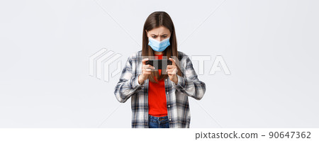 Different emotions, covid-19 pandemic, coronavirus self-quarantine and social distancing concept. Serious focused young girl in medical mask trying pass level in mobile game, tap screen and frowning 90647362