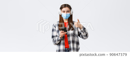 Social distancing, lifestyle on covid-19 outbreak, coronavirus concept. Cheerful pleased good-looking woman in medical mask, approve nice sound of wireless headphones, thumb-up, hold phone Social distancing, lifestyle on covid-19 outbreak, coronavirus concept. Cheerful pleased good-looking woman in medical mask, approve nice sound of wireless headphones, thumb-up, hold phone 90647379