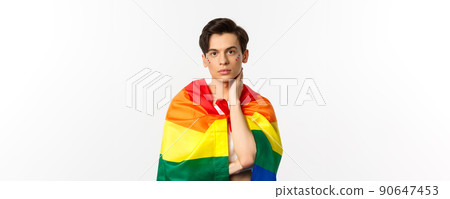 Waist-up shot of beautiful gay man with glitter on face, wrap himself with rainbow lgbt flag and touching face gently, looking at camera, white background Waist-up shot of beautiful gay man with glitter on face, wrap himself with rainbow lgbt flag and touching face gently, looking at camera, white background 90647453