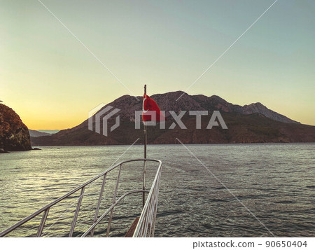 yacht in the open sea. sea with a sandy beach against the backdrop of a mountain. the turkish red flag is installed on the stern of the yacht yacht in the open sea. sea with a sandy beach against the backdrop of a mountain. the turkish red flag is installed on the stern of the yacht 90650404