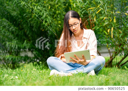 Young Vietnamese woman sitting on grass loan working on tablet outside in park Young Vietnamese woman sitting on grass loan working on tablet outside in park 90650525