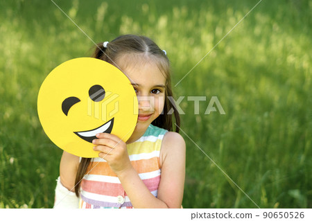 A beautiful attractive brunette girl holds a winking emoticon while closing one eye. Smile day world. Funny emoticon A beautiful attractive brunette girl holds a winking emoticon while closing one eye. Smile day world. Funny emoticon 90650526