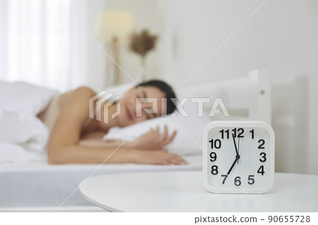 Alarm clock set for 7 AM on bedside table, with happy woman sleeping on bed in background Alarm clock set for 7 AM on bedside table, with happy woman sleeping on bed in background 90655728