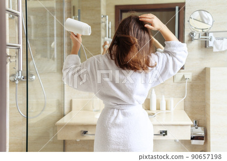 Back view of woman in a white robe blow dry hair by hair dryer standing and looking in front of the mirror in the bathroom 90657198