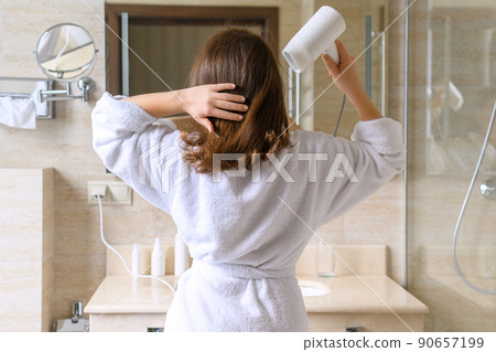 Back view of woman in a white robe blow dry hair by hair dryer standing and looking in front of the mirror in the bathroom 90657199