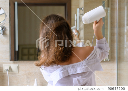 Back view of woman in a white nightgown blow dry hair by hair dryer standing and looking in front of the mirror in the bathroom 90657200