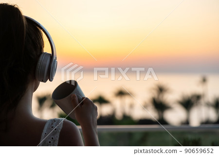 Rear view of woman with headphones and cup of tea looking on palm trees sea beach at sunset. Female relaxing listenning music. Rear view of woman with headphones and cup of tea looking on palm trees sea beach at sunset. Female relaxing listenning music. 90659652