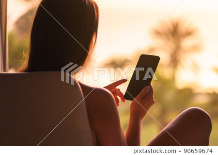 Rear view of woman looking at phone sitting on armchair near panoramic window opposite palm trees sea beach at sunset. 90659674