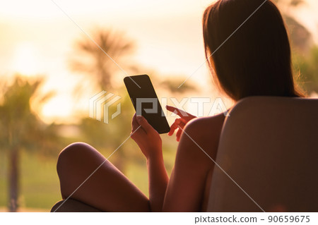 Rear view of woman looking at phone sitting on armchair near panoramic window opposite palm trees sea beach at sunset. 90659675