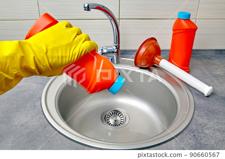 Hands in yellow rubber gloves hold a plunger and a pipe cleaner against the background of a kitchen sink 90660567