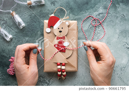 Hands in the process of wrapping a Christmas gift, decorated with a wooden Christmas tree toy in the form of a bear in a santa claus hat with a heart in his paws with the inscription xmas. Flat lay 90661795