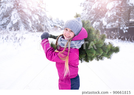 On a frosty winter sunny day, a happy girl 10-11 years in winter clothes carrying a Christmas tree home, to decorate for the holiday, smiling, looking at camera. Winter Holidays Eve. 90661815