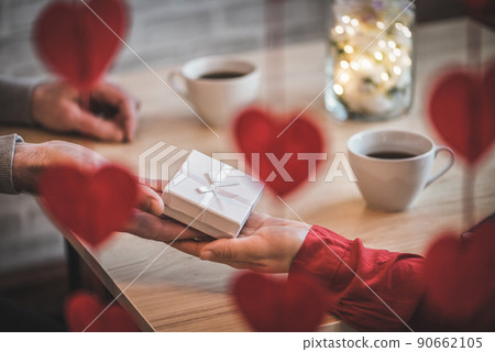 Valentines day. Close-up of woman and man celebrating in restaurant. Boyfriend giving small white gift box to girlfriend. View through a heart-shaped garland. love, romance, date Valentines day. Close-up of woman and man celebrating in restaurant. Boyfriend giving small white gift box to girlfriend. View through a heart-shaped garland. love, romance, date 90662105