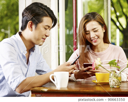 happy and smiling young asian couple using mobile phone while relaxing in coffee shop or tea house 90663350